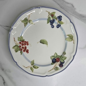 May include: White ceramic plate with a scalloped edge and a blue rim. The plate features a painted design of red currants, blueberries, and blackberries with green leaves. A light blue stripe encircles the center of the plate.