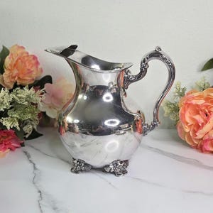 May include: A silver-colored pitcher with a curved handle and ornate detailing. The pitcher has a wide spout and sits on three decorative feet. The background includes pink and orange flowers, suggesting a vintage or antique aesthetic.