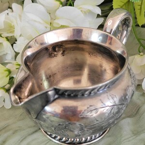 May include: A silver creamer with a floral design. The creamer has a rounded body and a small spout. The handle is curved and has a rounded end.