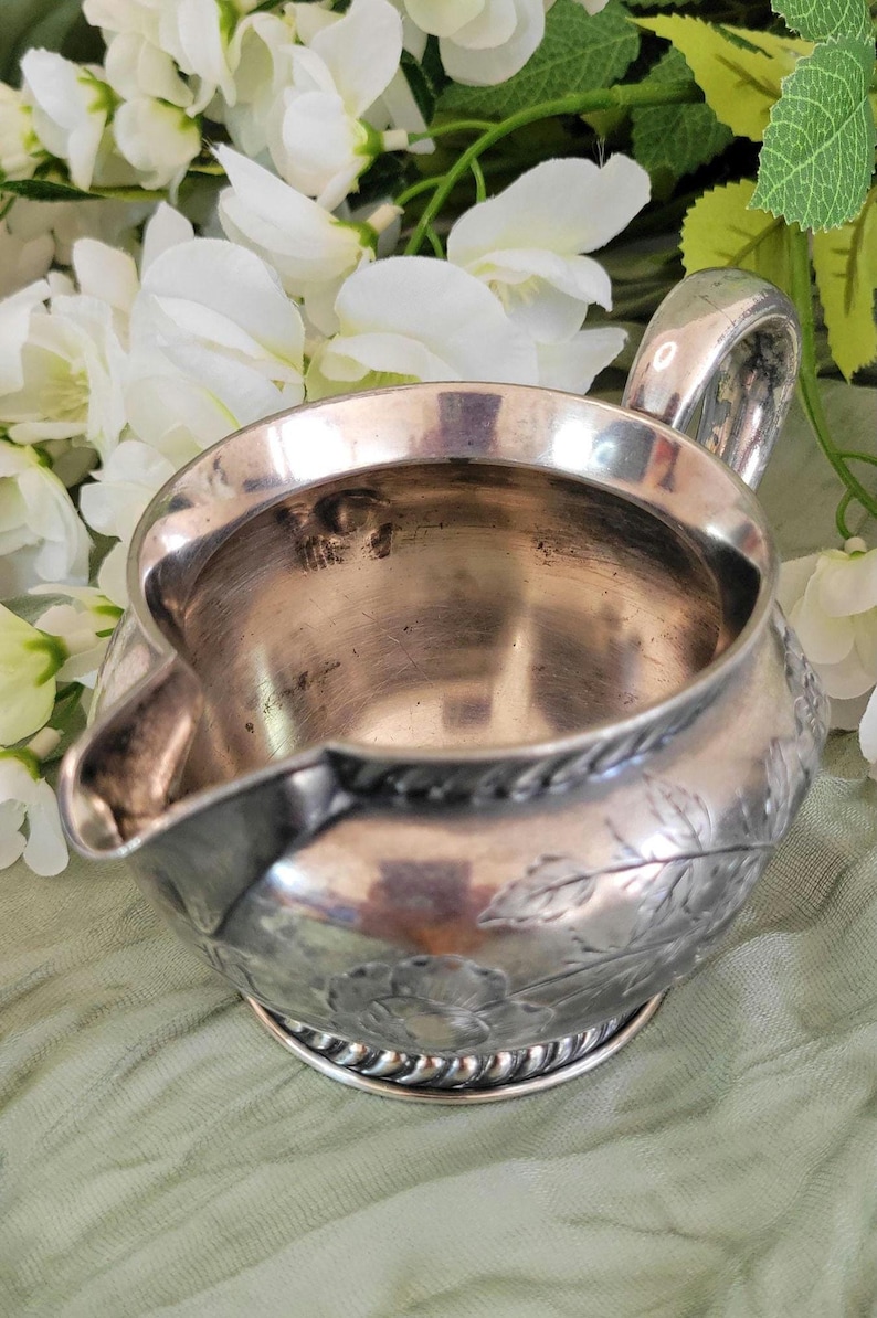 May include: A silver creamer with a floral design. The creamer has a rounded body and a small spout. The handle is curved and has a rounded end.