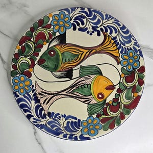 May include: A round, hand-painted ceramic plate featuring two stylized fish in the center. The fish are green, yellow, and orange, with black details. The plate is decorated with blue floral and leaf patterns, and measures approximately 10 inches in diameter.