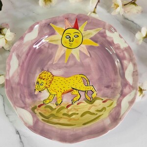 May include: A ceramic plate with a pink background and a yellow lion walking across a green field. The sun with a face is in the upper center of the plate.