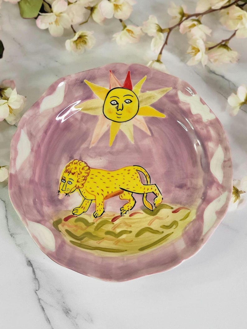 May include: A ceramic plate with a pink background and a yellow lion walking across a green field. The sun with a face is in the upper center of the plate.