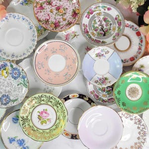 May include: A collection of vintage teacup saucers in various colors and floral patterns. The saucers are arranged on a white surface, with some featuring gold trim and intricate designs. Some saucers have a pink, blue, green, and white color scheme.
