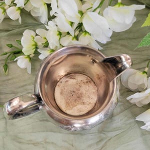 May include: Silver creamer with a decorative design and a handle. The creamer is empty and has a worn, aged look.