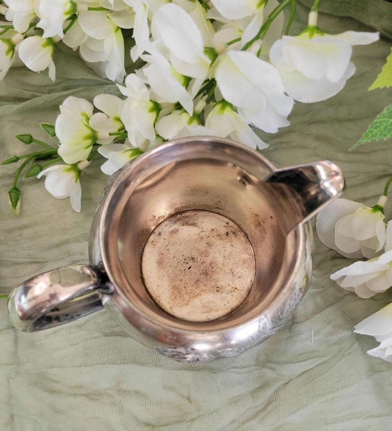 May include: Silver creamer with a decorative design and a handle. The creamer is empty and has a worn, aged look.
