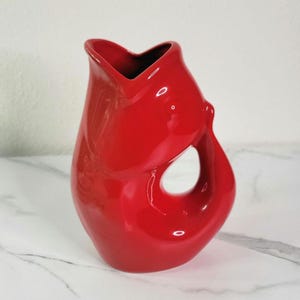 May include: A glossy red ceramic vase with a unique, organic shape. The vase features a large, circular opening and a flared top. The interior is dark, contrasting with the bright red exterior. A decorative piece for home decor.
