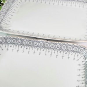 May include: Two rectangular white serving platters with a gray and gold decorative border. The platters have a scalloped edge and are placed on a white surface. Lemons and greenery are in the background.