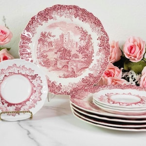 May include: Set of pink and white vintage china plates with a floral pattern and a castle scene. The plates are arranged on a white surface.