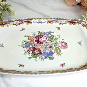 May include: Rectangular white ceramic serving tray with a floral design. The tray features a central bouquet of pink, purple, and blue flowers. The border has a gold and burgundy pattern with small floral accents. A decorative piece for serving.