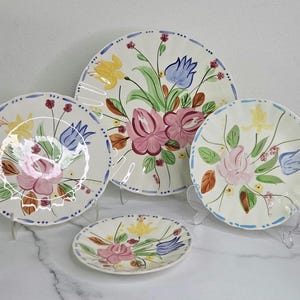 May include: A set of four ceramic plates with floral designs. The plates feature a cream-colored background with pink, yellow, and blue flowers. Each plate has a blue dotted border. The plates are displayed on clear stands.