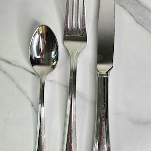 May include: A three-piece stainless steel flatware set consisting of a spoon, a four-tined fork, and a dinner knife. Each piece features a polished, reflective finish and a handle with vertical grooved detailing that tapers toward the base.