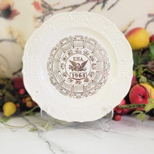 May include: White ceramic plate with a gold embossed design of the 1965 calendar for the United States. The plate has a scalloped edge and a raised design around the rim.
