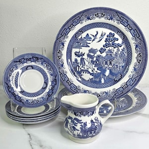 May include: A collection of blue and white Willow pattern china, including plates, saucers, and a creamer. The plates feature a detailed landscape design with pagodas, bridges, and birds. The creamer has a handle and a similar blue and white pattern.