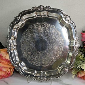 May include: A silver-colored, ornate serving tray with a scalloped edge. The tray features a detailed, symmetrical pattern in the center and along the rim. The tray is supported by a clear stand, showcasing its elegant design.