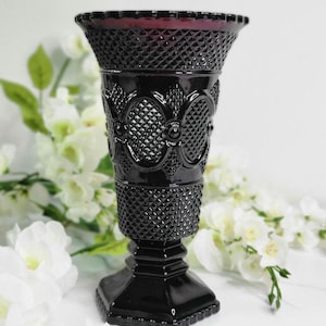 May include: A black glass vase with a diamond and oval pattern. The vase has a flared top with a ruffled edge and a square base. The vase is approximately 10 inches tall.