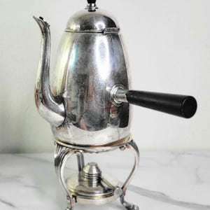 May include: A silver-colored coffee pot with a black handle and a black knob on top. The pot sits on a decorative stand with curved legs. The spout is curved and elegant. A classic kitchenware item.