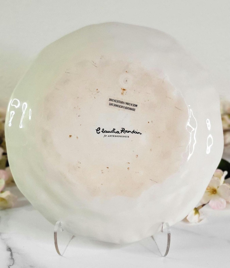 May include: White ceramic plate with a slightly rippled surface. The plate has a black inscription that reads "Claudia Rankin for Anthropologie" and "DISHWASHER & MICROWAVE SAFE MADE IN CHINA/FABRIQUE EN CHINE".