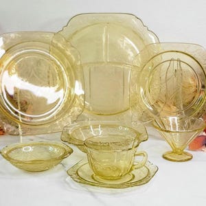 May include: A collection of vintage yellow glass tableware, including plates, bowls, a cup and saucer, and a serving dish. The pieces have a floral pattern and are arranged on a white surface.
