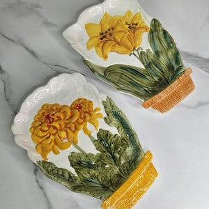 May include: Two white, scalloped-edge decorative plates. Each plate features a vibrant yellow flower design with green leaves and a brown pot base. The plates are likely ceramic and are set against a marble background.