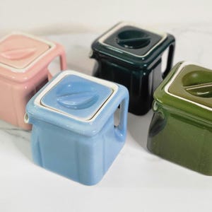 May include: Four ceramic teapots in different colors: pink, blue, green, and dark green. Each teapot has a square shape with a lid and a spout.