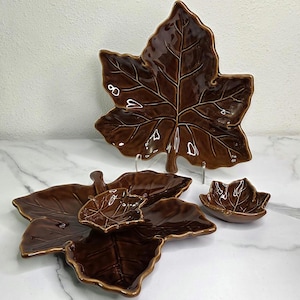 May include: A set of brown ceramic leaf-shaped serving dishes. The set includes a large platter, several smaller plates, and a small bowl. The dishes have a glossy finish and are arranged on a white surface.