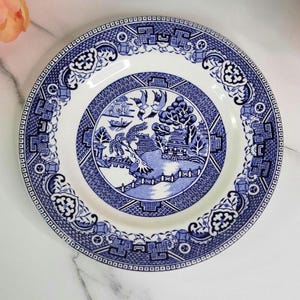 May include: A round, white and blue ceramic plate with a classic Willow pattern design. The plate features a central scene with a bridge, birds, and a pagoda, surrounded by a detailed border. The plate is approximately 10 inches in diameter.