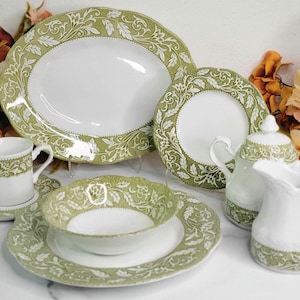 May include: A white porcelain tea set with a green floral design. The set includes a large oval platter, a round plate, a bowl, a cup and saucer, a teapot, and a creamer. The edges of the pieces have a scalloped design.