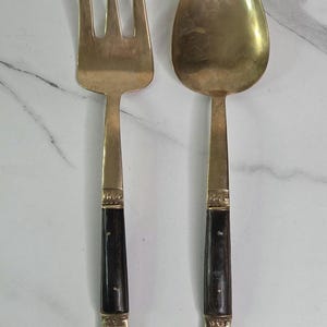May include: A vintage serving fork and spoon set. The utensils are made of gold-colored metal with dark, possibly ebony, handles. The handles are adorned with ornate, decorative details at the base. The fork has three tines.