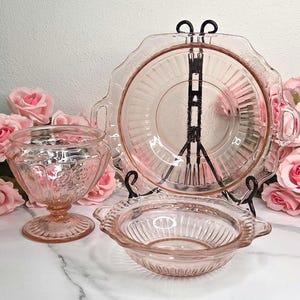 May include: A set of three pieces of pink depression glass including a footed bowl, a shallow handled bowl, and a large handled serving plate. Each piece features a radial ribbed pattern and embossed floral details on the translucent glass surface.