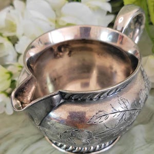 May include: A silver creamer with a floral design. The creamer has a rounded body and a small spout. The design features leaves and flowers.