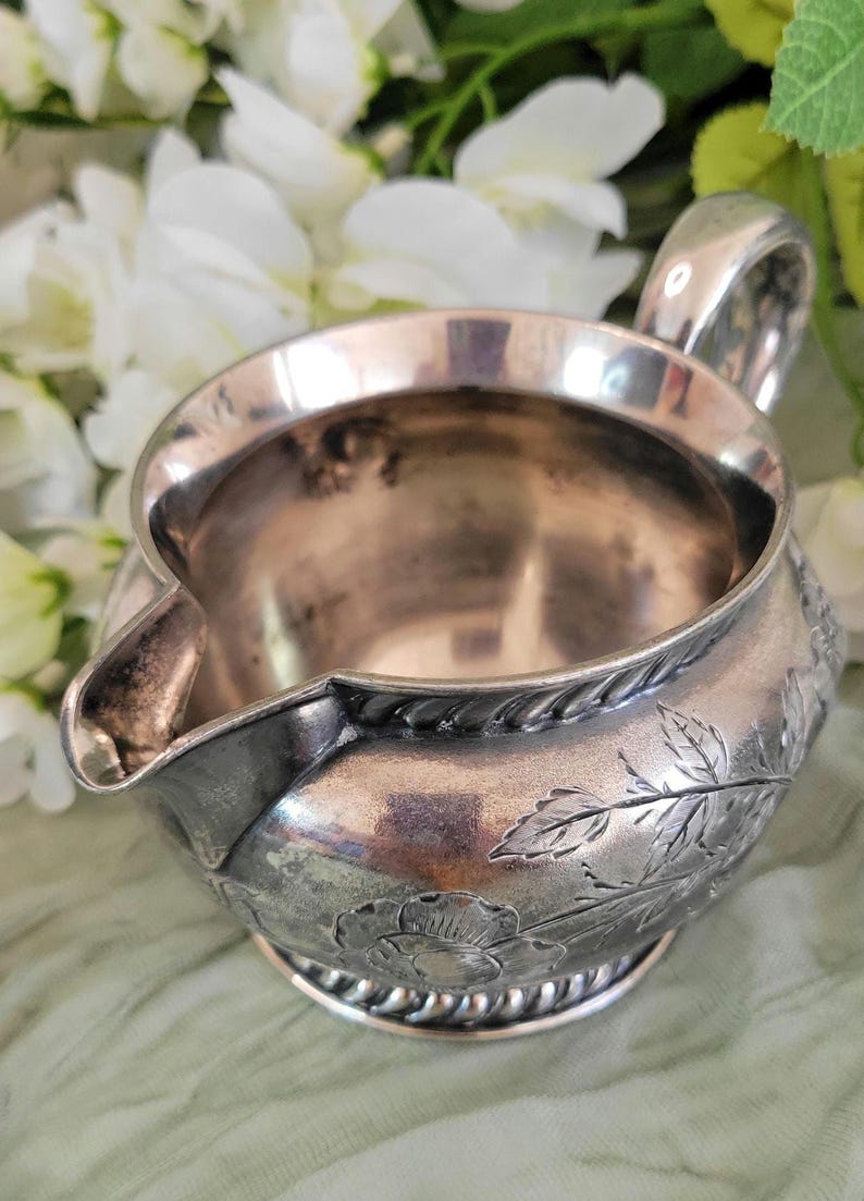 May include: A silver creamer with a floral design. The creamer has a rounded body and a small spout. The design features leaves and flowers.