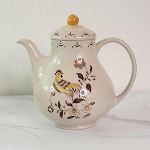 May include: A cream-colored ceramic teapot with a brown and yellow floral design featuring a rooster. The teapot has a lid with a small knob on top.