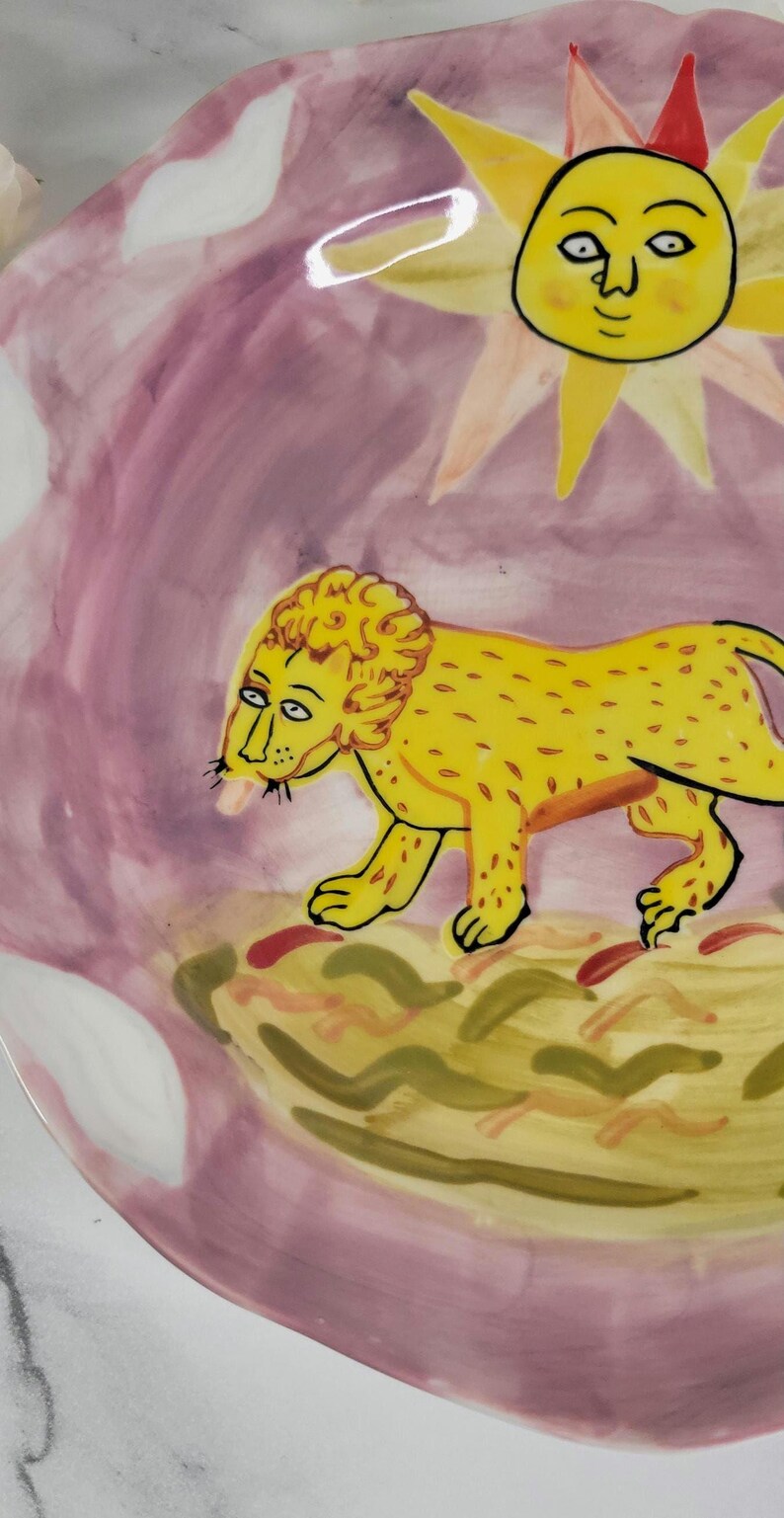 May include: A pink ceramic plate with a yellow lion and a yellow sun with a face painted on it.