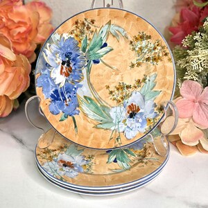 May include: Set of three decorative plates with a floral design. The plates feature a light orange background with blue and white flowers and green leaves. The plates have a blue trim.