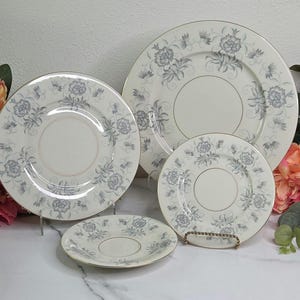 May include: A set of four white porcelain plates with a floral pattern in shades of gray and gold trim. The set includes a dinner plate, salad plate, saucer, and a smaller plate, perfect for a formal dining setting.