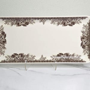 May include: Rectangular serving platter with a cream-colored background and a brown floral border design. The platter has two handles and is supported by a clear stand. The floral design includes leaves and flowers.