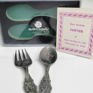 May include: A set of Early American pewter silverware, including a fork and spoon with ornate handles. The spoon features a detailed design. The set comes with a box containing two green spoons and a card from Web Silver Co. Inc.