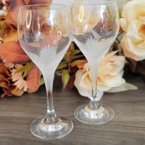 May include: Two clear glass wine glasses with a frosted design. The glasses have a tall, slender stem and a wide bowl. The glasses are sitting on a wooden surface.