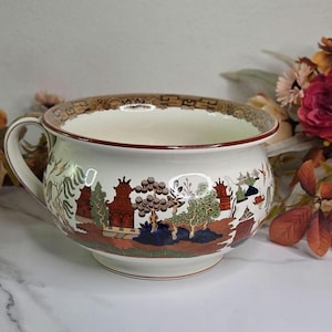 May include: A white ceramic chamber pot with a handle, featuring a detailed landscape scene in shades of red, green, and blue. The rim has a brown and gold patterned border. Decorative flowers surround the pot.