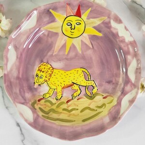 May include: A ceramic plate with a pink background and a yellow lion with a sun with a face above it.