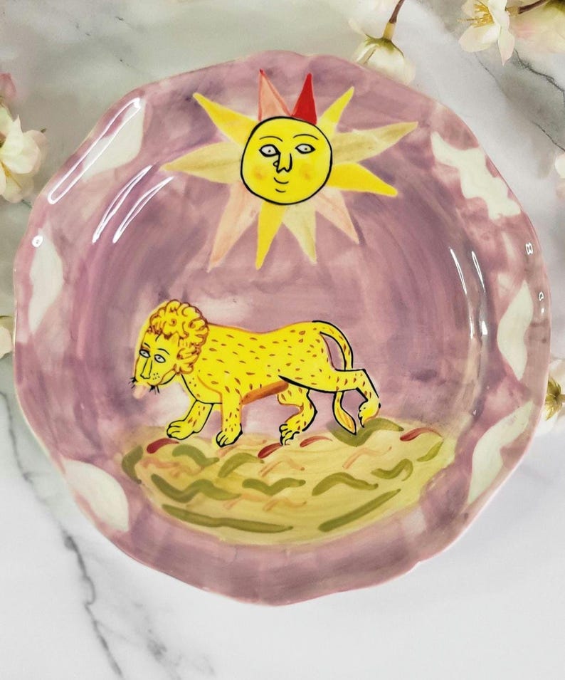 May include: A ceramic plate with a pink background and a yellow lion with a sun with a face above it.