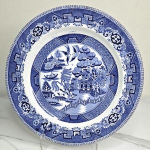 May include: A round, white and blue ceramic plate with a classic Willow pattern design. The plate features a detailed landscape scene with a bridge, trees, and birds. The rim has a geometric and floral border. The plate is displayed on a clear stand.