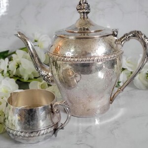 May include: A silver teapot and creamer set with ornate details. The teapot has a rounded body with a hammered finish and a lid with a finial. The creamer has a similar hammered finish and a small handle.