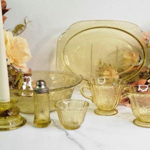 May include: A collection of vintage yellow glassware, including a serving tray, bowl, cups, creamer, and a small dish. A candle in a yellow candlestick holder and a salt shaker are also present. The glassware is decorated with a floral pattern.