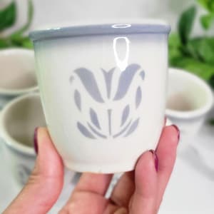 May include: A white ceramic cup with a light blue floral design and a light blue rim. The cup is held in a hand, with other similar cups visible in the background. The cup is approximately 3 inches tall.