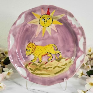 May include: A pink ceramic plate with a white rim features a hand-painted design of a yellow lion walking across a green and brown landscape. A yellow sun with a smiling face is painted in the upper center of the plate.
