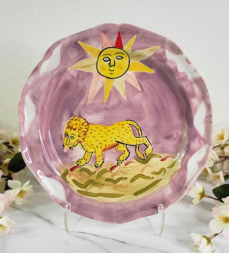 May include: A pink ceramic plate with a white rim features a hand-painted design of a yellow lion walking across a green and brown landscape. A yellow sun with a smiling face is painted in the upper center of the plate.