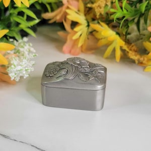 May include: A small, silver, rectangular box with an ornate floral design on the lid. The box is made of pewter and has a smooth, polished finish.