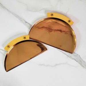 May include: Two half-circle, copper-colored trays with a yellow, curved handle. Each handle has three star-shaped cutouts. The trays are on a white marble surface. The trays are likely decorative or serving pieces.